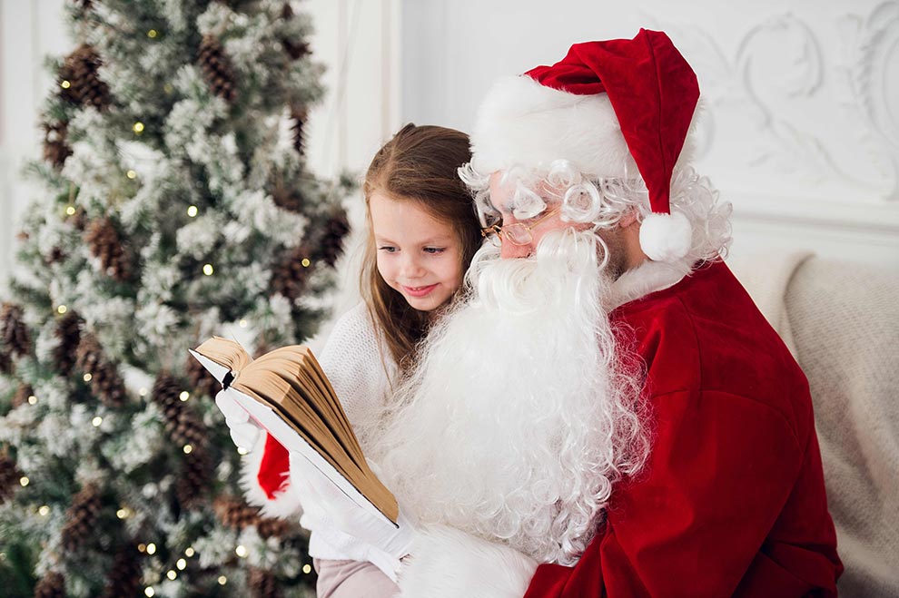 Santa Claus reads a book to a girl sitting on his lap.