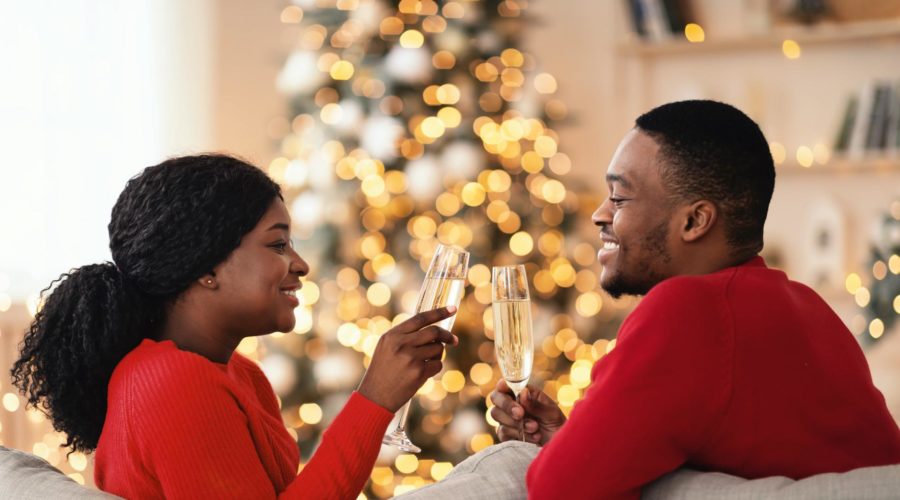 Happy young couple in love clink glasses with champagne, sit on sofa with tree with garlands in living room.