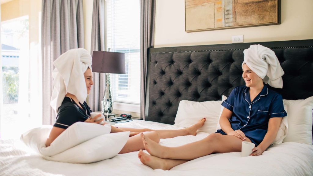 Two women with bath towels over their hair sitting and talking on a king bed in an Encore Resort residence during a girls’ getaway.