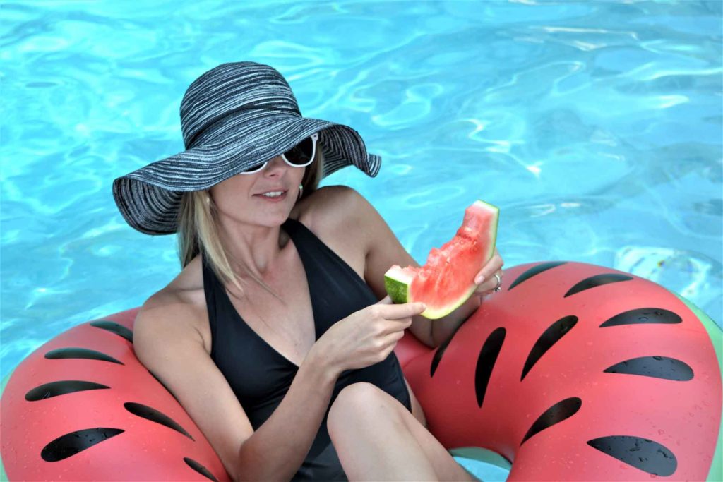 Woman in a swimsuit and sun hat eating a slice of watermelon while floating on a tube decorated like a watermelon in a swimming pool.