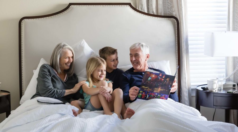 Happy grandparents reading to two young grandchildren in a large king bed.