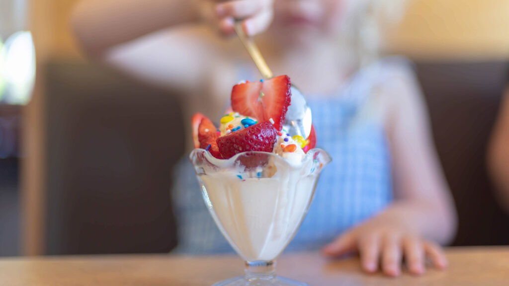 Closeup of ice cream sundae with strawberries and sprinkles with child's hand holding spoon